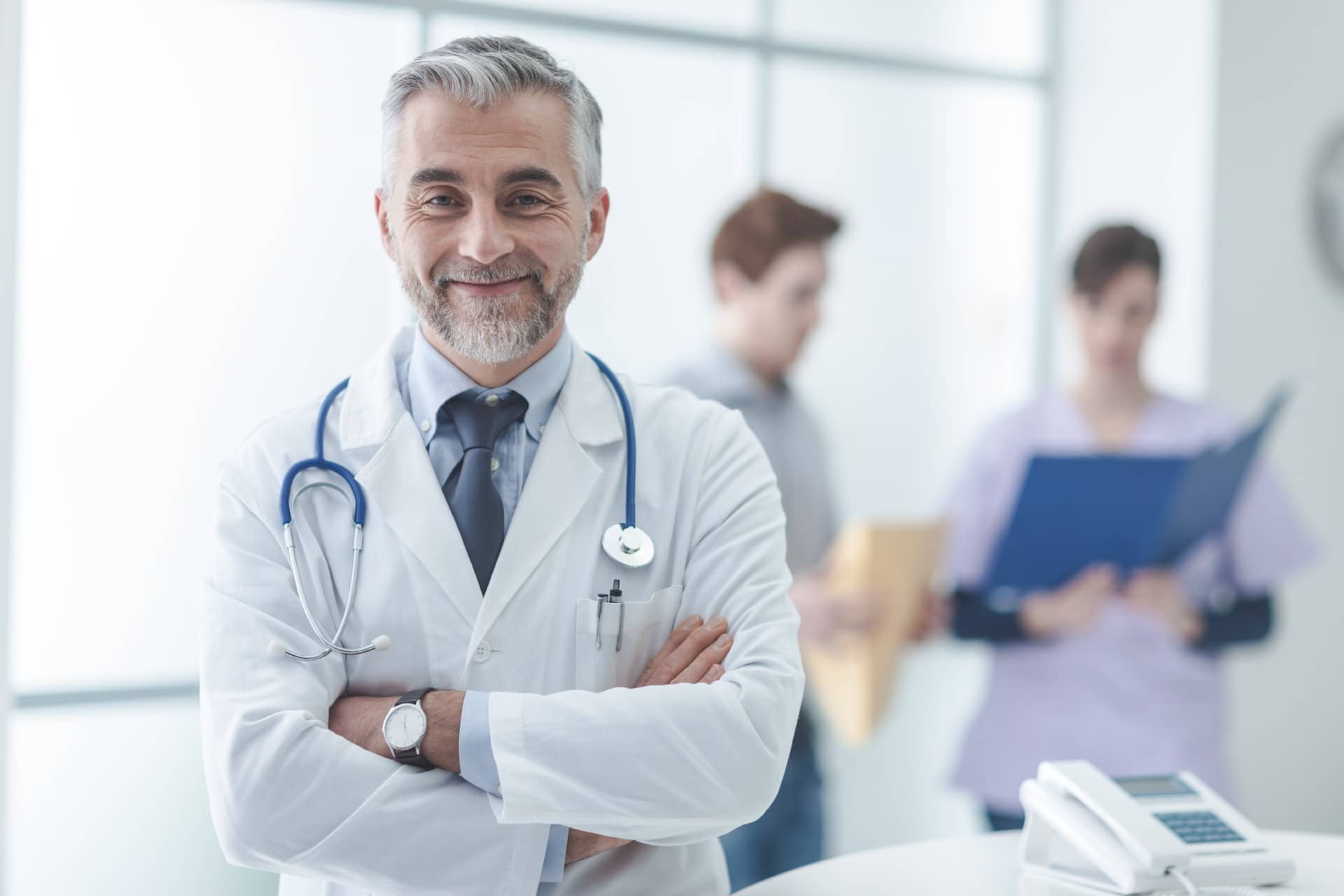 a doctor smiling at you with his arms crosses with 2 people in teh background looking at a folder